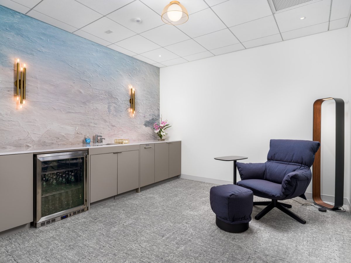 JAMS LA wellness room — beverage cabinet, navy swivel chair with ottoman, hand-troweled cream-to-blue gradient plaster wall, brass tube sconces.