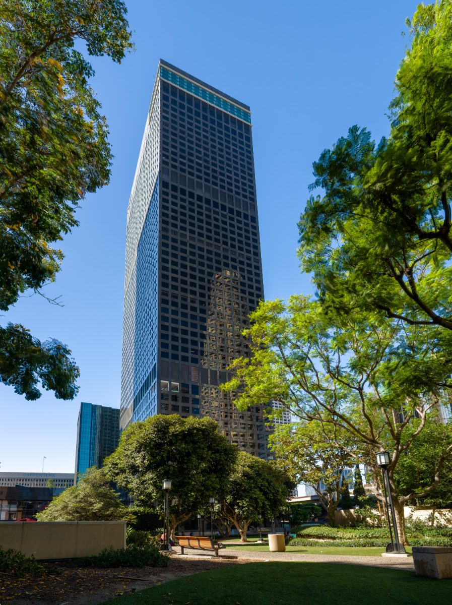 Vertical exterior of City National Plaza, downtown LA — dark glass-and-steel tower housing JAMS LA, framed by trees in the plaza garden, cyan crown overhead.