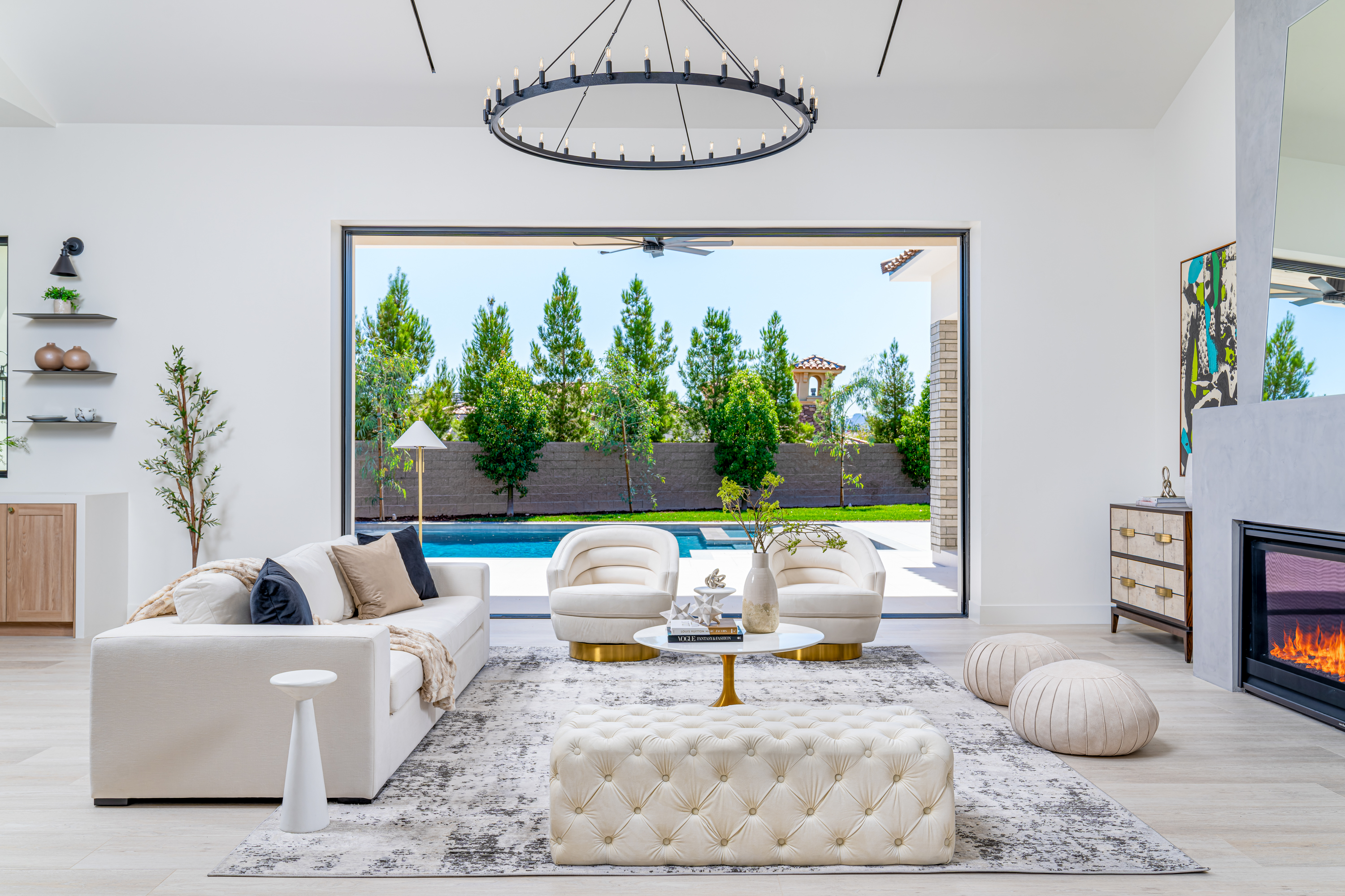 Living Room at a Stately Vegas-luxury indoor-outdoor home in Las Vegas (16 CHC) — wagon-wheel chandelier, tufted ottoman, pleated barrel chairs, pool view.