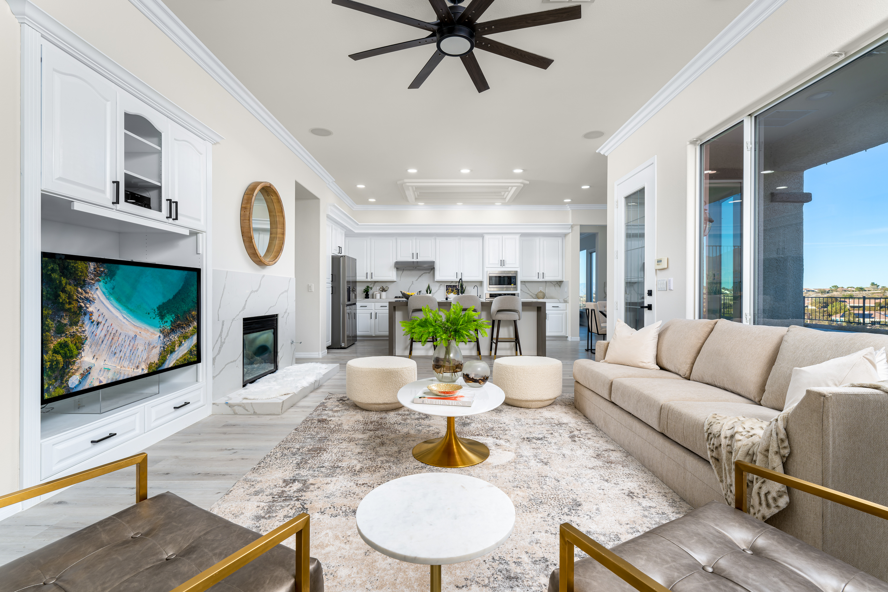 Living Room at a Stately casual coastal elegance home in Las Vegas (1952 OSS) — beige sectional, dark wood ceiling fan, gold-leg side tables, bouclé poufs.
