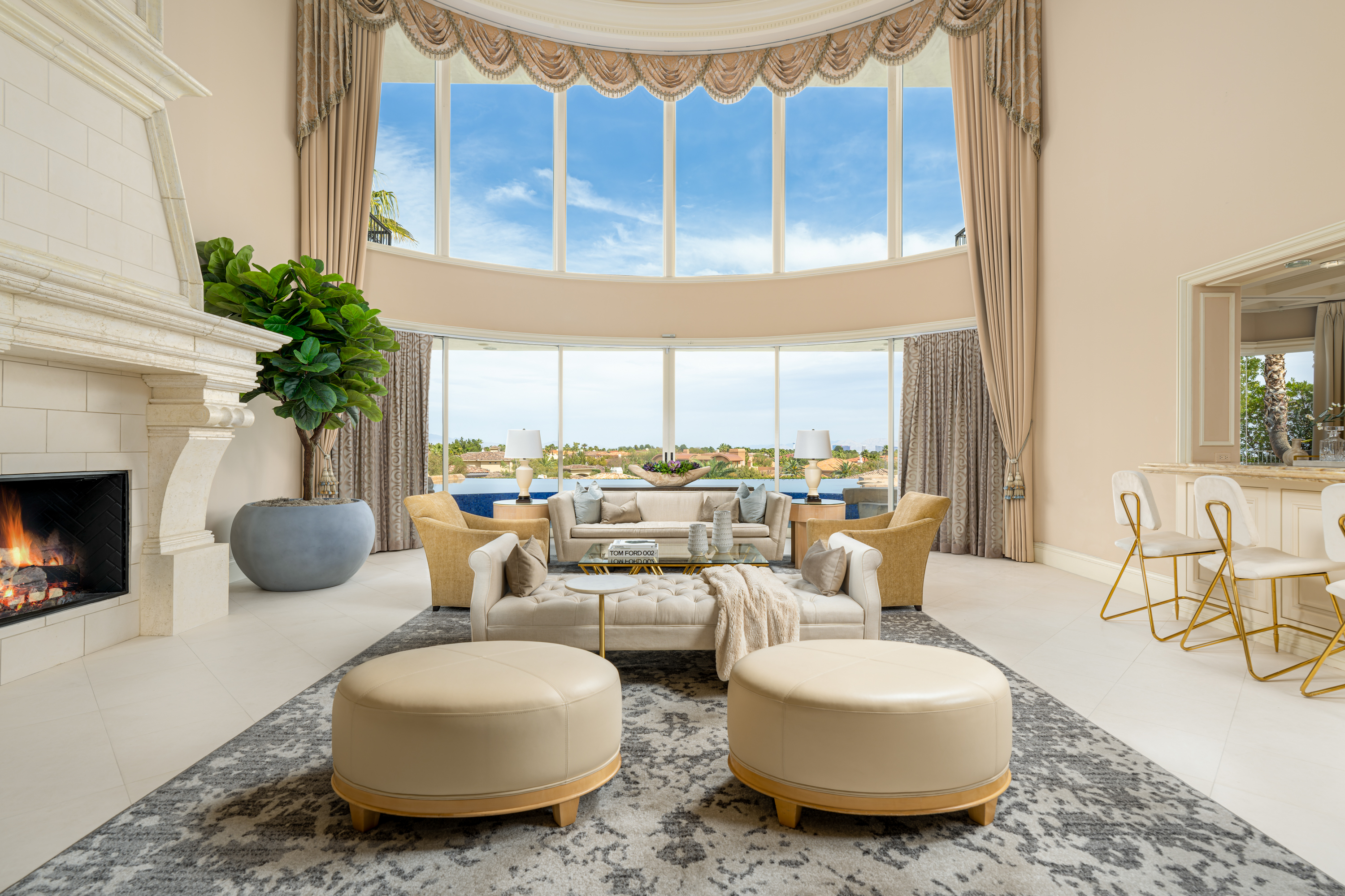 Living Room at a Stately glam estate home in Las Vegas (5114 SHD) — two-story curtains, paired gold-base ottomans, marble fireplace, panoramic view.