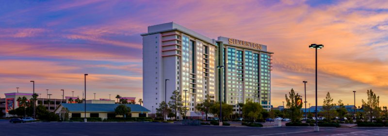 Silverton Casino Lodge at dawn — wide panoramic of the Las Vegas hotel tower with rooftop sign, Bass Pro Shops wing, pink-accent parking deck, peach cirrus sky.
