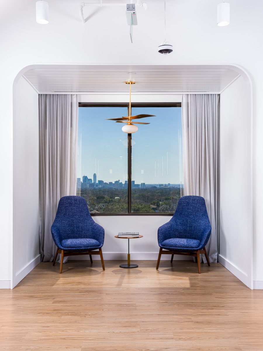 JAMS Dallas window-alcove nook — two blue tweed chairs flanking a pedestal table under a brass leaf chandelier, downtown Dallas skyline beyond at Sherry Lane Place.
