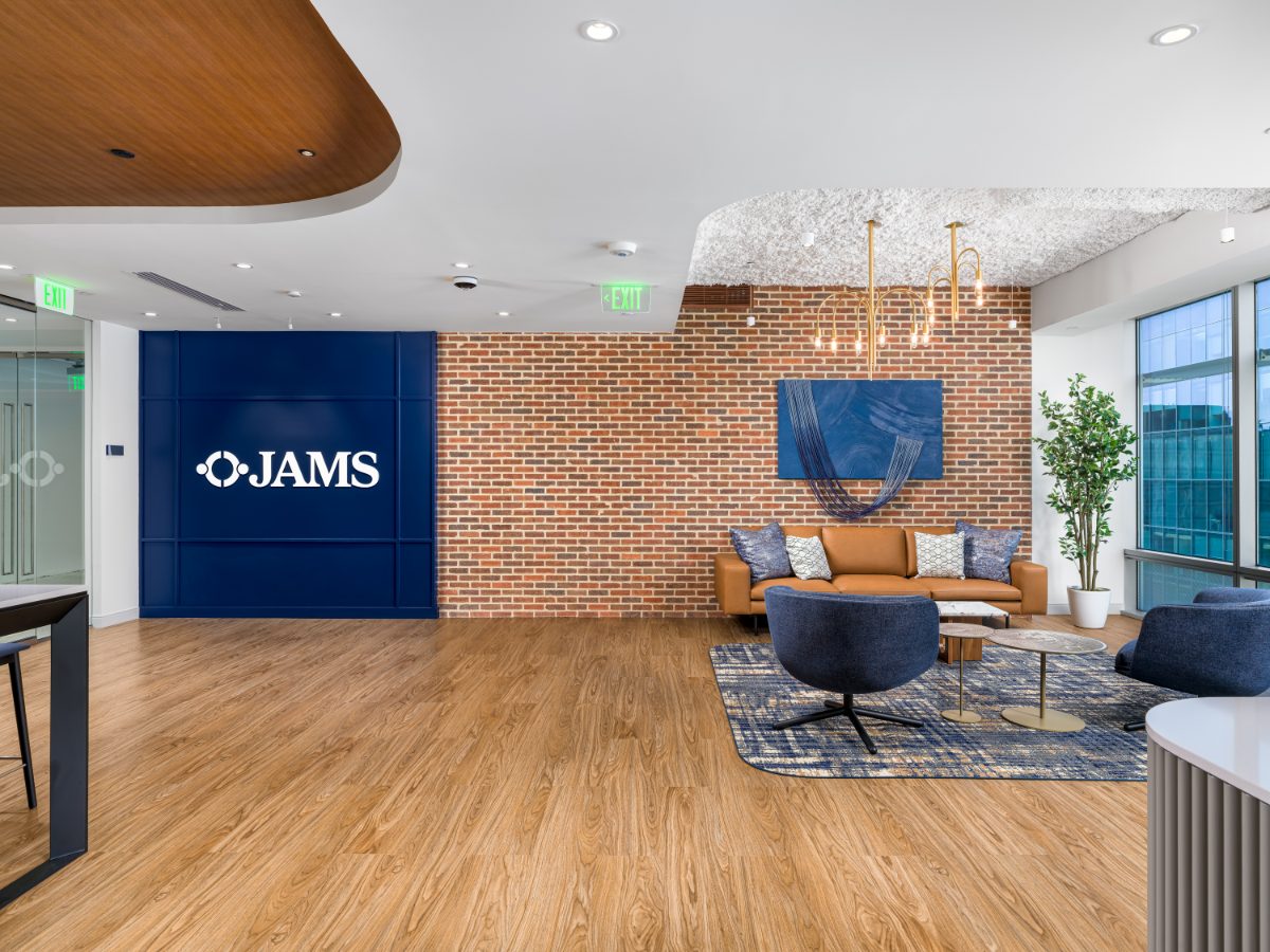JAMS DC reception wide-angle at 1050 K Street NW — navy JAMS brand wall, red brick accent wall, brass arched chandelier, tan leather sofa, blond plank floor.