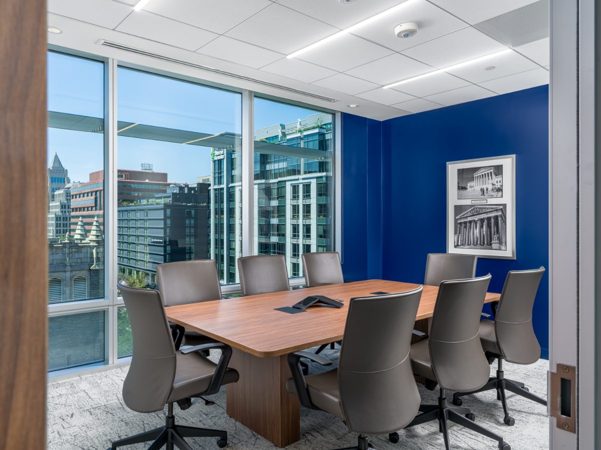 JAMS Washington, D.C. conference room shot from the doorway — walnut table, grey leather chairs, blue wall with Supreme Court prints, K Street skyline window.