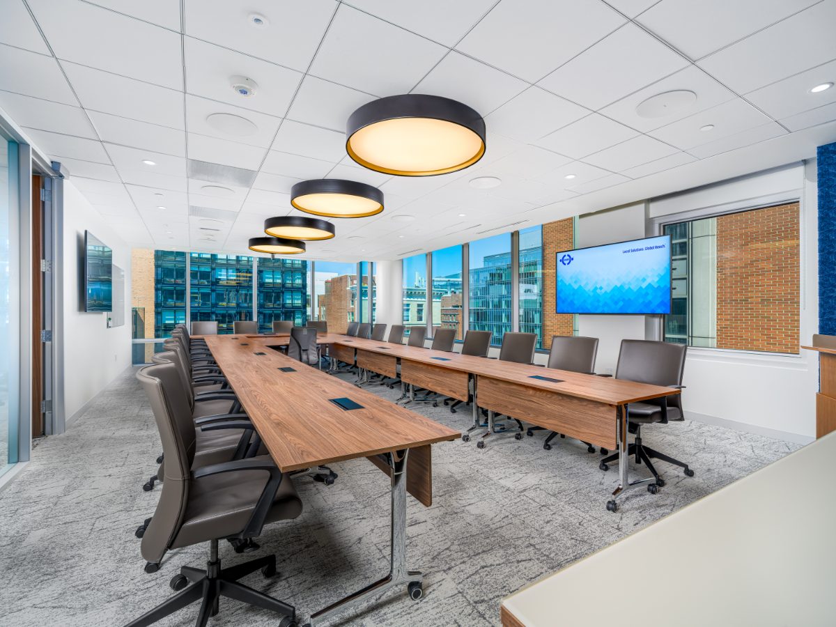 Grand boardroom at JAMS Washington, D.C. — two long walnut tables, twenty grey leather chairs, four drum pendants, JAMS displays, and K Street windows.