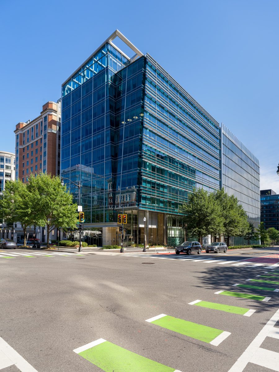 1050 K Street NW shot vertical — modern glass office tower's crystalline corner prow climbing into a clear blue sky, with green bike-lane bars in the foreground.