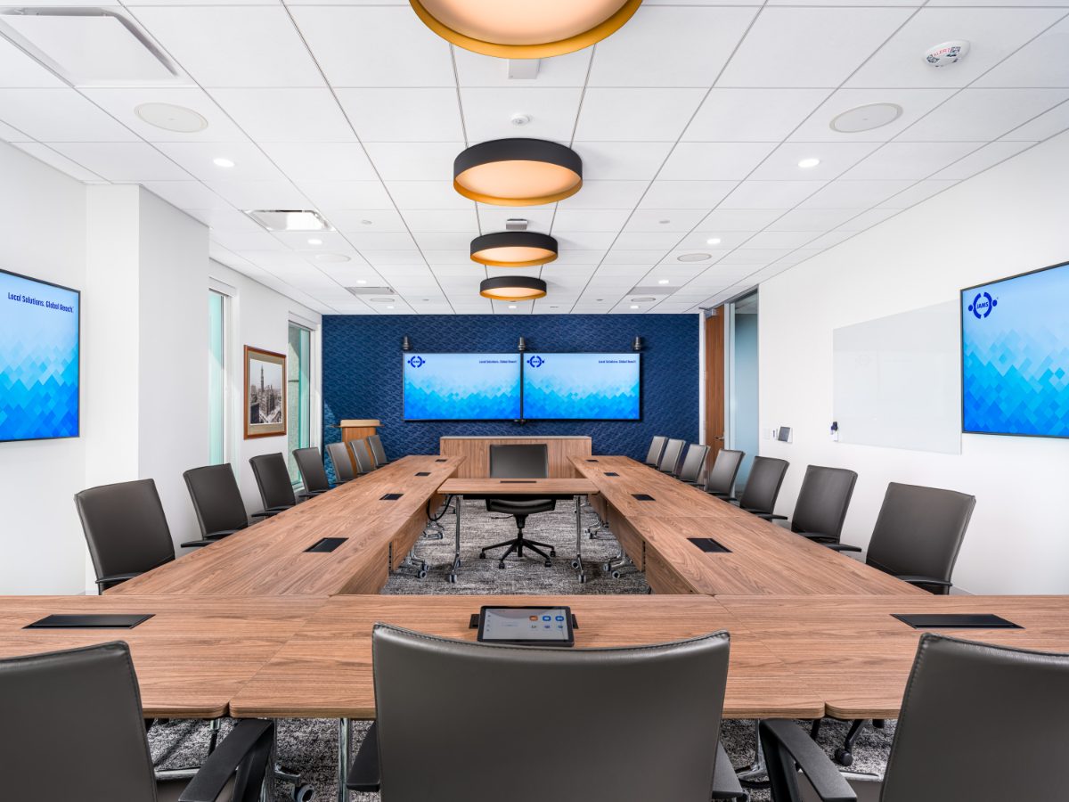 JAMS Detroit grand boardroom down the length — walnut U-shape, grey leather chairs, drum pendants in perspective, three JAMS displays on a navy back wall.