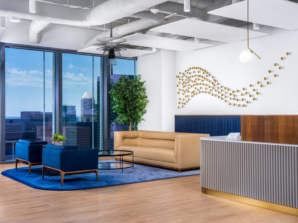 JAMS Houston reception lounge — cognac sofa, navy chairs on a blue rug, curved fluted desk, brass-orb wave sculpture, and downtown Houston skyline windows.