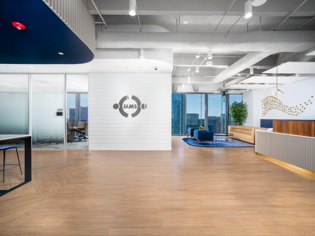 Wide-angle lobby at JAMS Houston — freestanding white shiplap wall with JAMS logo, glass conference room, reception lounge, and herringbone hardwood floor.