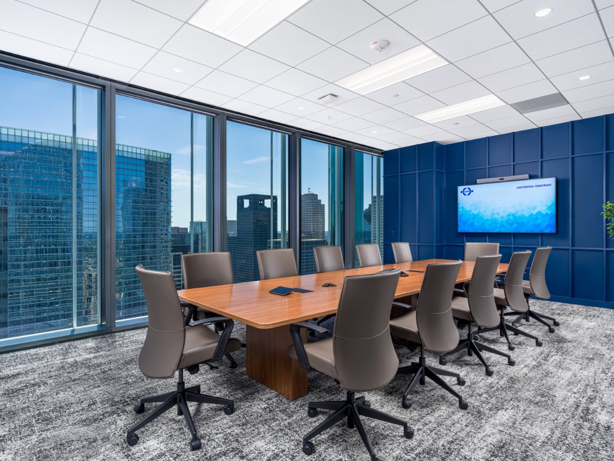 Panoramic conference room at JAMS Houston — walnut table, grey leather chairs, cobalt panel wall with JAMS display, and two skyline window walls wrapping the corner.