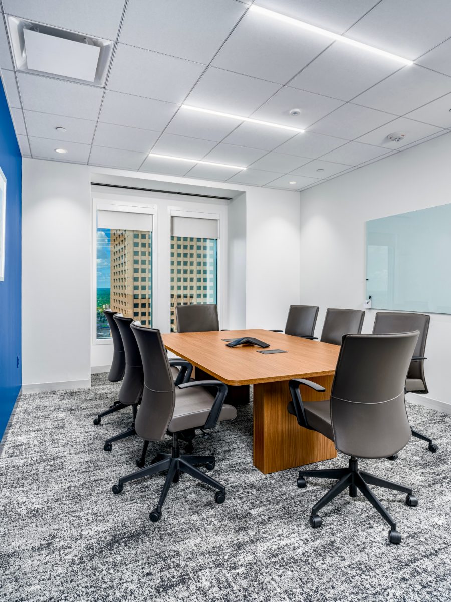 JAMS MN small mediation room — honey-wood table on plinth, grey leather chairs, cobalt accent edge, narrow window view of a neighboring Minneapolis tower.