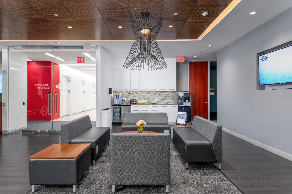 JAMS IAC reception lounge — grey modular seating, dramatic sculptural black wire-cage chandelier overhead, small mini café behind, walnut perforated ceiling.