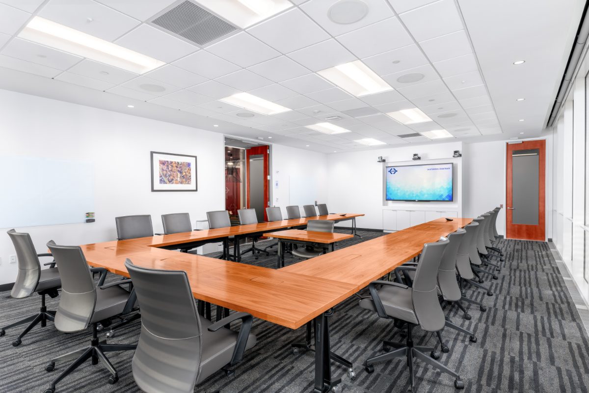 Boardroom 7 at JAMS NY 16th Floor — U-shaped cherry tables, grey leather chairs, twin ceiling cameras flanking a JAMS display, framed aerial art, red doors, window wall.