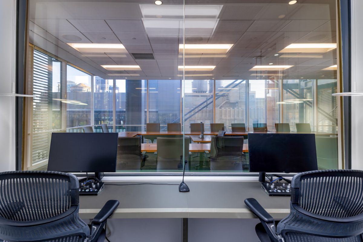 Translation booth at JAMS NY 16th Floor — two Aeron chairs, dual Dell monitors and gooseneck mic, tinted glass onto Boardroom 7 and Midtown skyline.