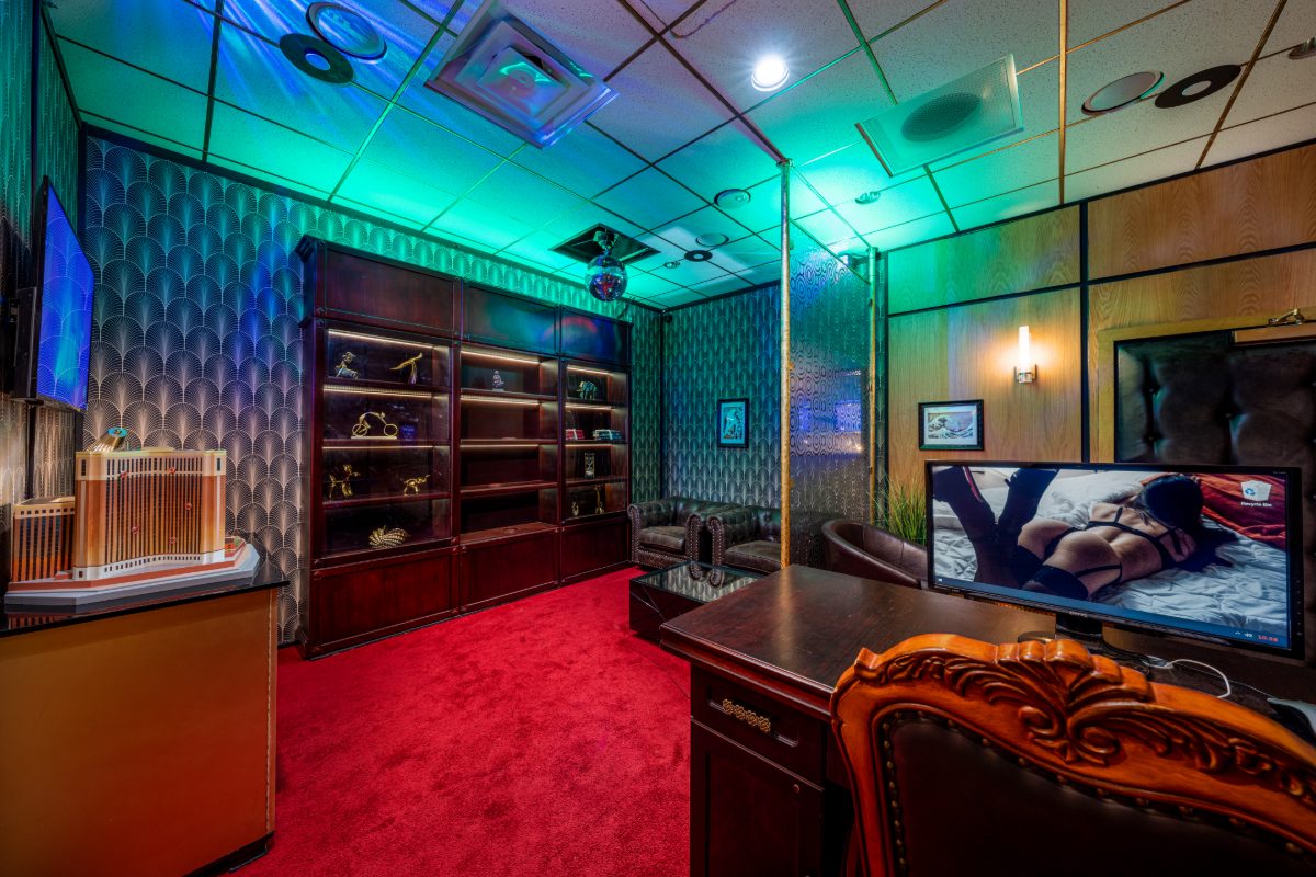 Wide-angle interior of PanIQ's Casino Heist escape room — Art Deco fan-pattern wallpaper, mahogany trophy cabinet with gold figurines on LED-lit shelves, scale model of a casino bu