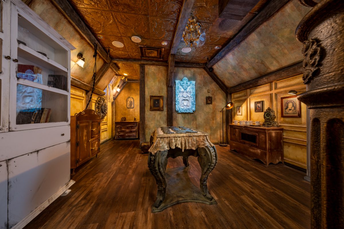 Interior of The Haunted Manor escape room at PanIQ Las Vegas, styled as the Vesper Castle parlor — embossed pressed-tin ceiling in deep amber, small crystal chandelier, mottled age